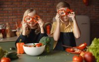 Funny boy and girl wearing apron looking through slices of pepper while preparing healthy vegetarian salad together at table with ingredients and bowls