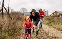 A happy family enjoys a bonding moment outdoors with a child learning to ride a bicycle on a fall day.