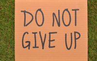 Cardboard sign reading 'Do Not Give Up' placed on green turf, symbolizing perseverance.